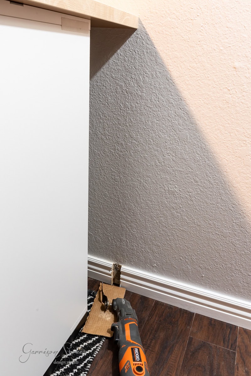 A multitool lies on a wooden floor next to a wall with a hole cut into it, perfectly suiting the needs of a DIY home office for two. The tool is partially resting on cardboard and blends seamlessly into the black-and-white patterned rug beside the white cabinet.