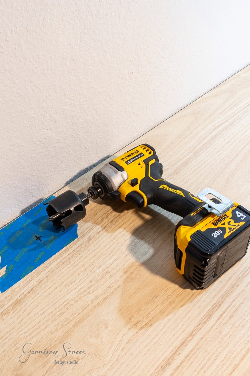 A yellow cordless power drill with a hole saw bit attached rests on a wooden surface, suggesting the start of a DIY home office for two. Blue painter’s tape with a marked “X” is on the wood, set against a plain, light-colored wall background.