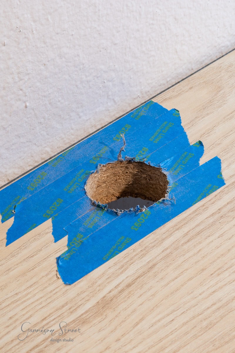 A close-up of a wooden surface with a hole cut out for a DIY home office for two. The area around the hole is covered with blue painter's tape to prevent splintering, revealing the rough edge of the wood beneath. A white wall looms in the background.