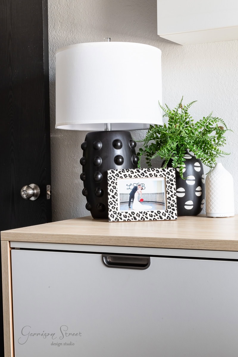 A modern tabletop in a DIY home office for two features a white lamp, black and white decorative vases, a small plant, and a leopard-print photo frame on a light wood and gray cabinet. A black door is partially visible in the background.