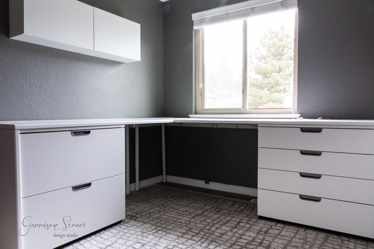 A minimalist room features gray walls and a white L-shaped desk for a seamless diy home office for two. The gray carpet complements the white cabinets with black handles, while the window offers a tranquil view of a tree outside.