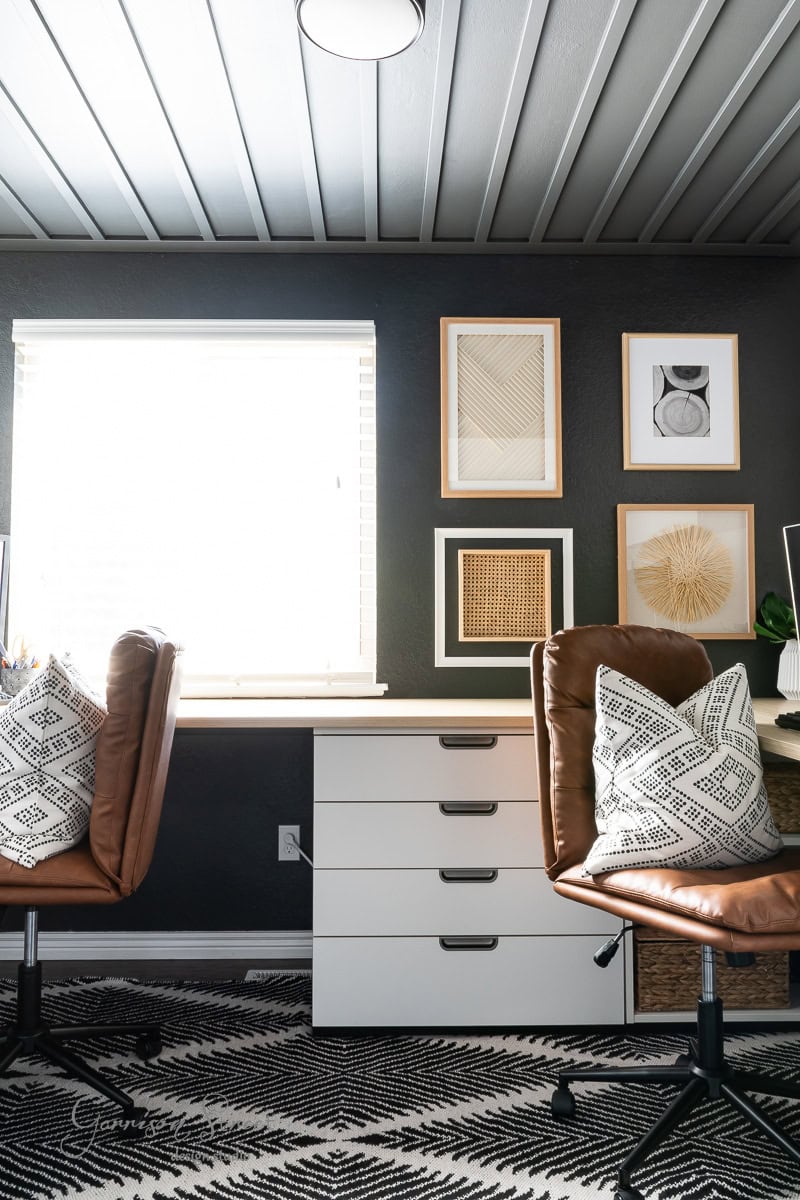 A DIY home office for two, this modern space boasts brown leather chairs with patterned cushions and a white desk with drawers. A gallery wall of framed art adds charm, while a left-side window filters natural light over dark gray walls and a patterned rug.