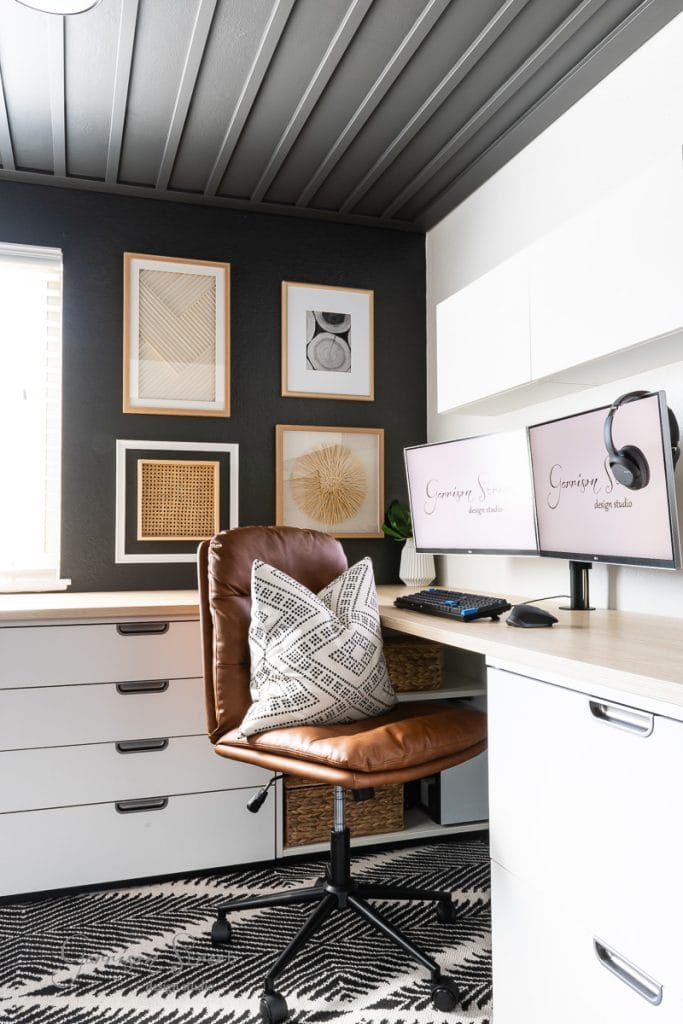 A modern DIY home office for two features dual computer monitors on a wooden desk, accompanied by a brown leather swivel chair and a black-and-white patterned rug. Framed abstract art adorns the walls, while a window invites natural light to enhance this inspiring workspace.