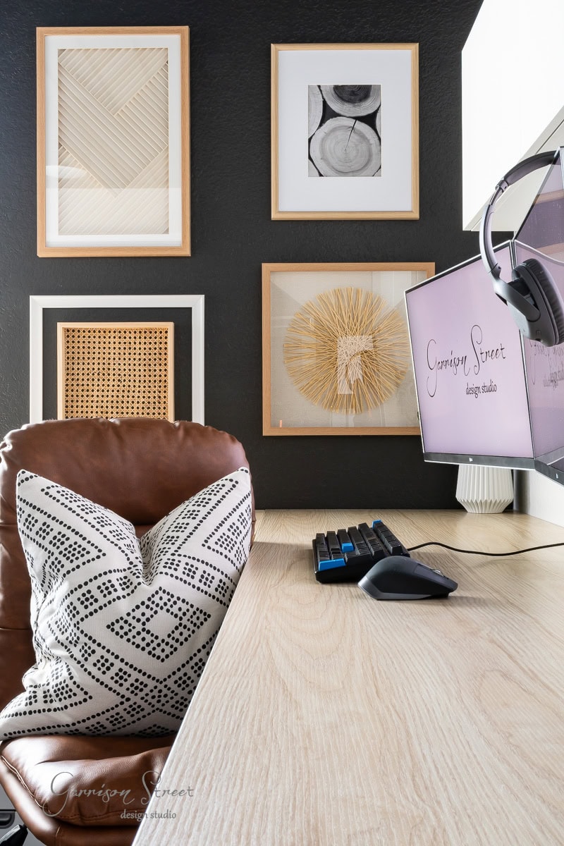 A modern DIY home office for two features a wooden desk and a leather chair with a patterned pillow. A computer monitor displays "Garrison Street Design Studio," while abstract artworks adorn the dark wall, creating an inspiring space for creativity and collaboration.