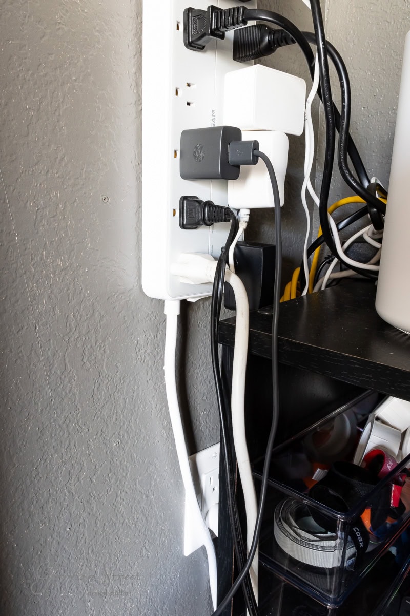 A power strip attached to a gray wall is filled with various black and white plugs and cables, efficiently serving this DIY home office for two. Nearby, a black shelf holds assorted items, including cables and a roll of tape, enhancing the workspace's organized aesthetic.