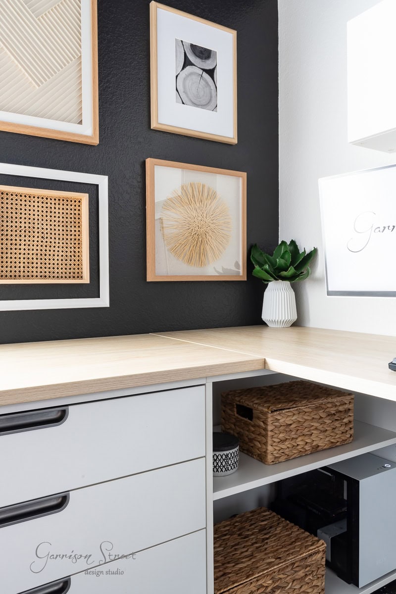 A modern DIY home office for two features a wooden desk nestled in a corner with a chic black accent wall. Framed abstract art and a small plant adorn the space, while woven baskets are neatly stored on white shelves beneath the desk.