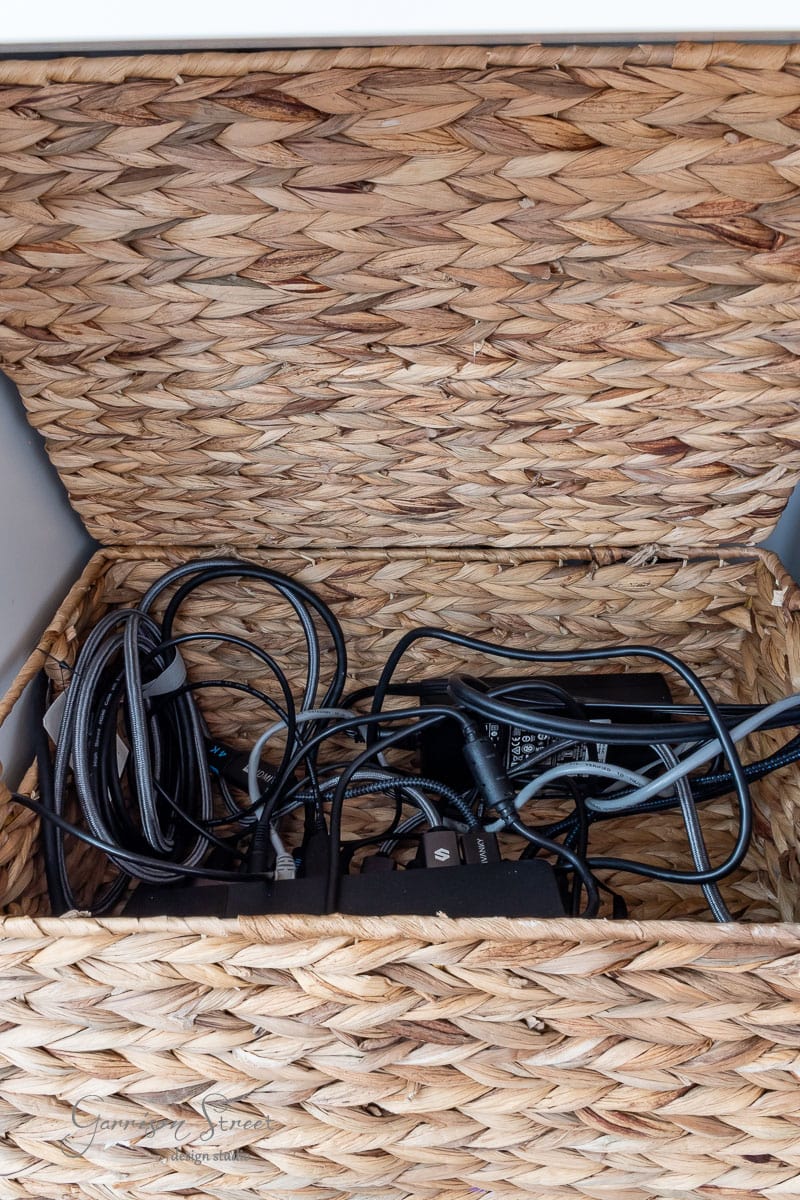 A woven basket with an open lid holds a tangled collection of black cables and chargers, perfect for organizing your DIY home office for two. Made of natural fibers, it sits elegantly on a neutral surface.