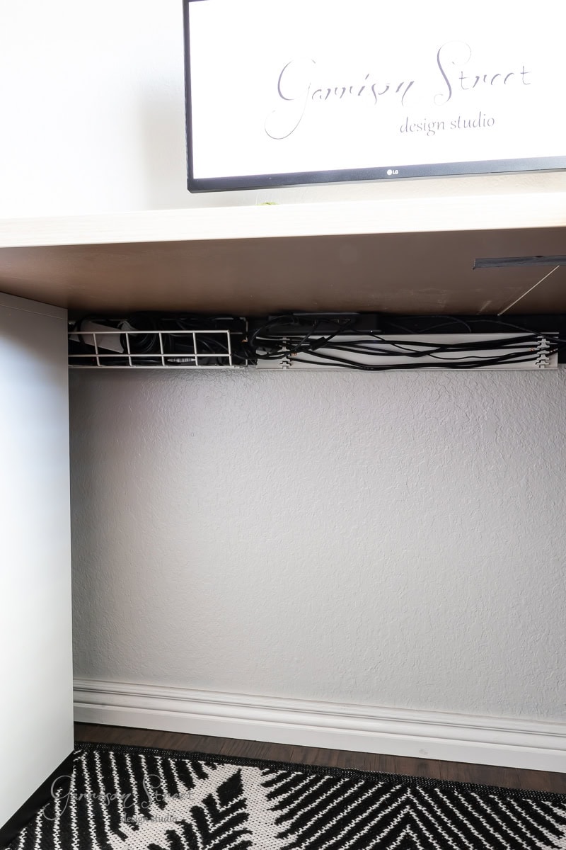 Under a desk, a cable management system neatly organizes various cables in this DIY home office for two. A portion of a monitor is visible at the top with the text "Garrison Street design studio." The floor features a black and white geometric patterned rug.