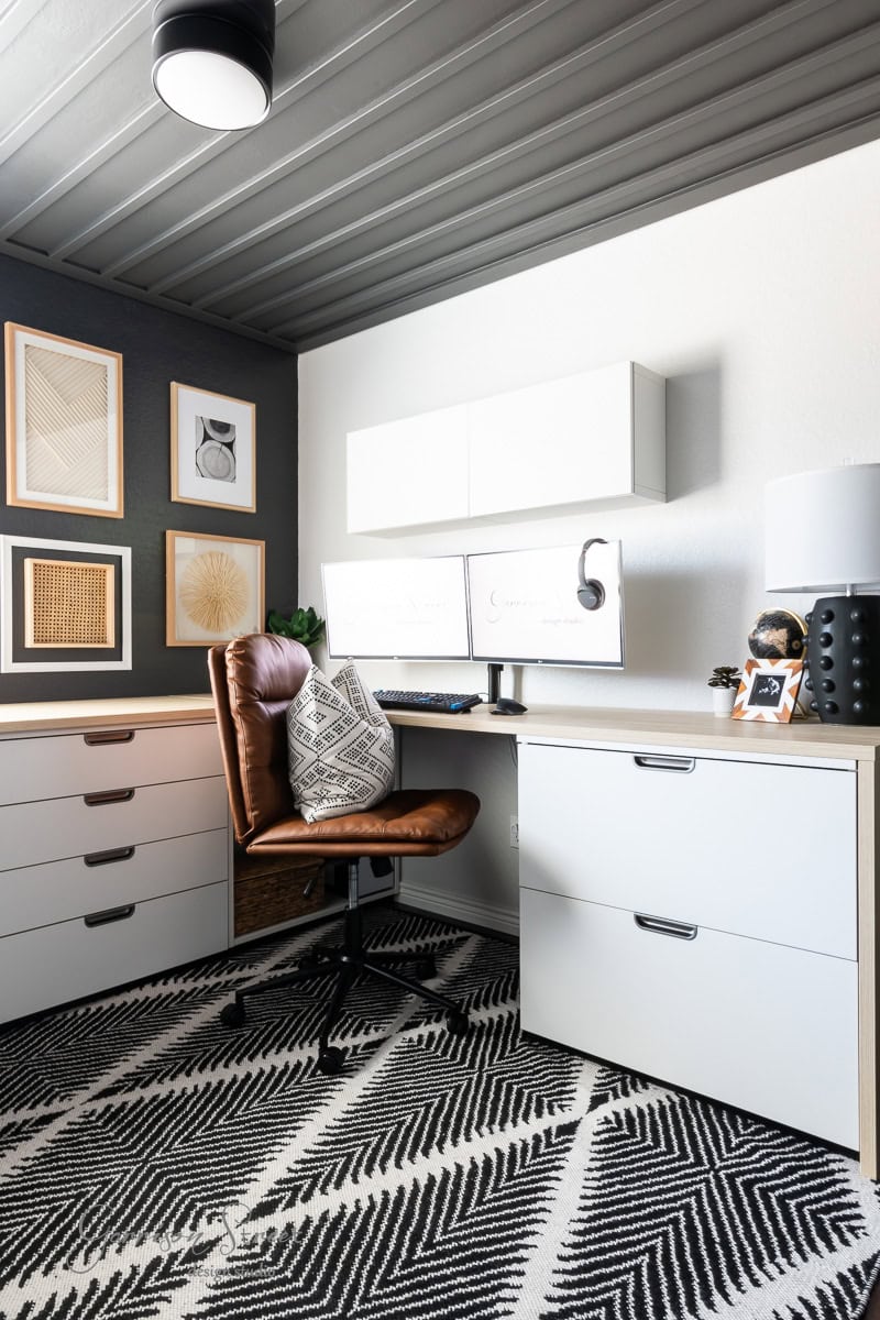 A modern DIY home office for two features a brown leather chair at a desk with dual monitors. White cabinets and a black accent wall with framed art add style. A patterned rug adorns the floor, creating a neatly organized and well-lit workspace perfect for productivity.