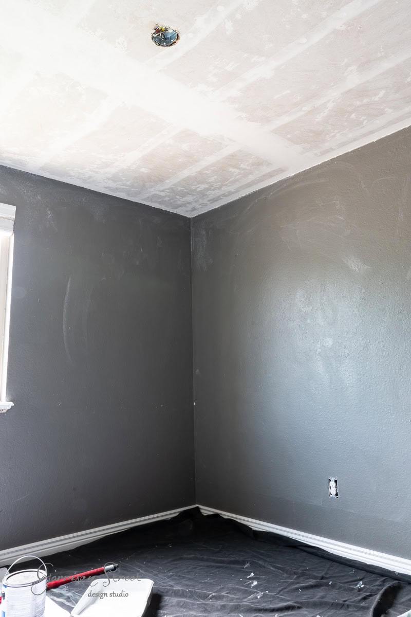 A room with dark gray walls is being transformed into a DIY home office for two. The unfinished ceiling with visible drywall adds a raw charm, while the window partially illuminates the space. Black protective cloth covers the floor, and painting supplies are neatly arranged in the corner.