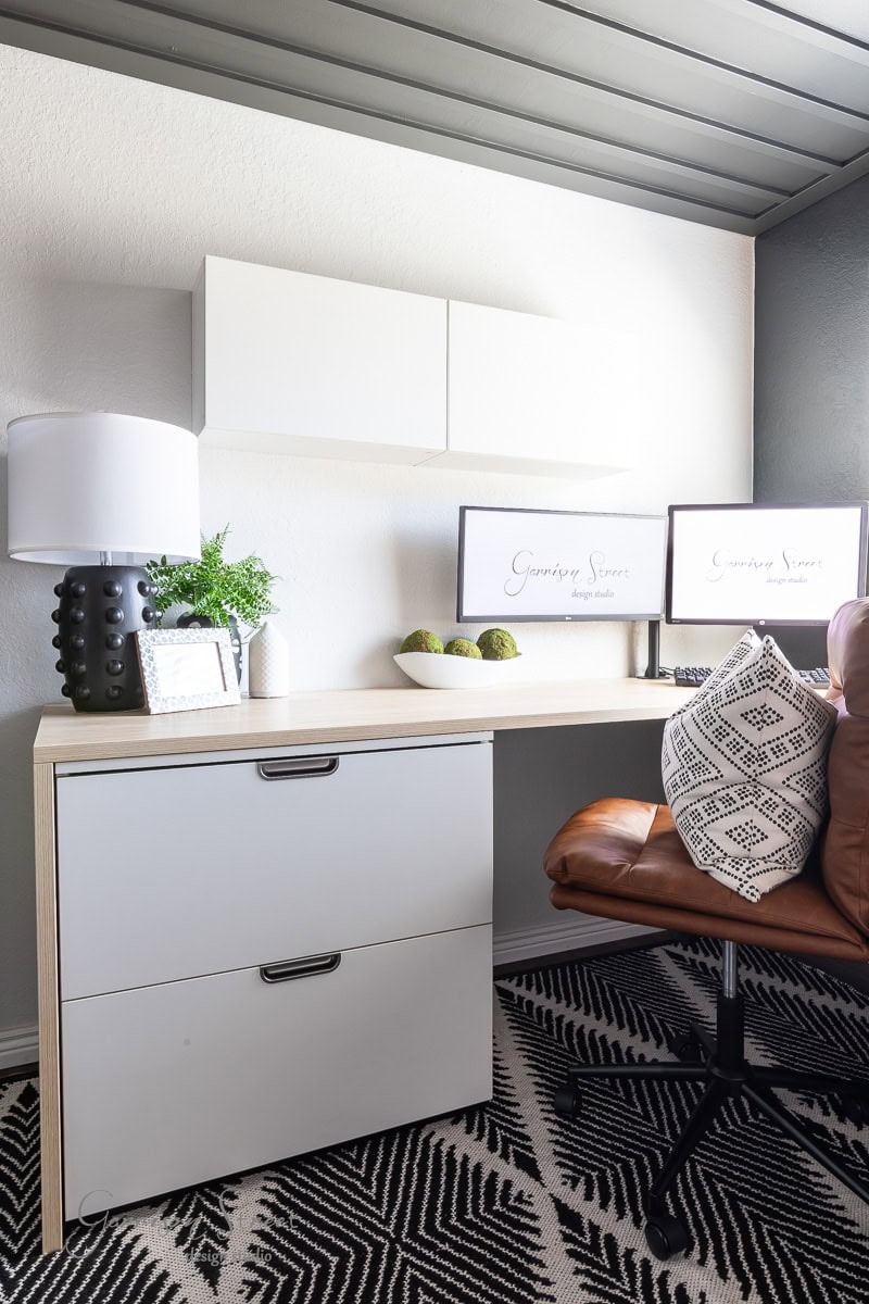 A modern DIY home office for two features a brown leather swivel chair, a wooden desk with white drawers, and dual monitors displaying "Gardenia Spaces." Decorative items include a lamp, framed photo, and plants. The room is tied together with a geometric black-and-white rug.