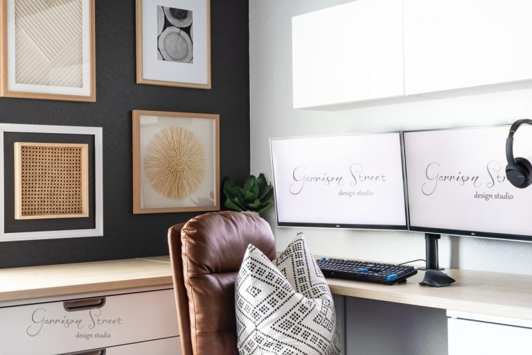 A modern DIY home office for two features a brown leather chair, black and white geometric pillow, dual monitors displaying "Garrison Street Design Studio," a keyboard, mouse, and wall art with framed abstract designs on contrasting dark and white walls.