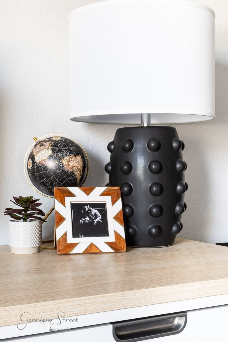 A wooden table in a DIY home office for two holds a spiky black lamp with a white shade, a small black globe with gold highlights, a framed black-and-white photo, and a potted succulent. The bottom left corner has the text "Garrison Street Design Studio.