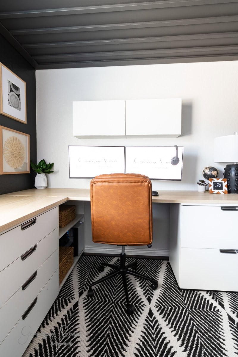 A modern DIY home office with a brown leather chair facing a white desk, perfect for two. The wall features minimalist decor and white cabinets above dual monitors. A black and white patterned rug covers the floor, while a small plant and lamp add charm to the workspace.