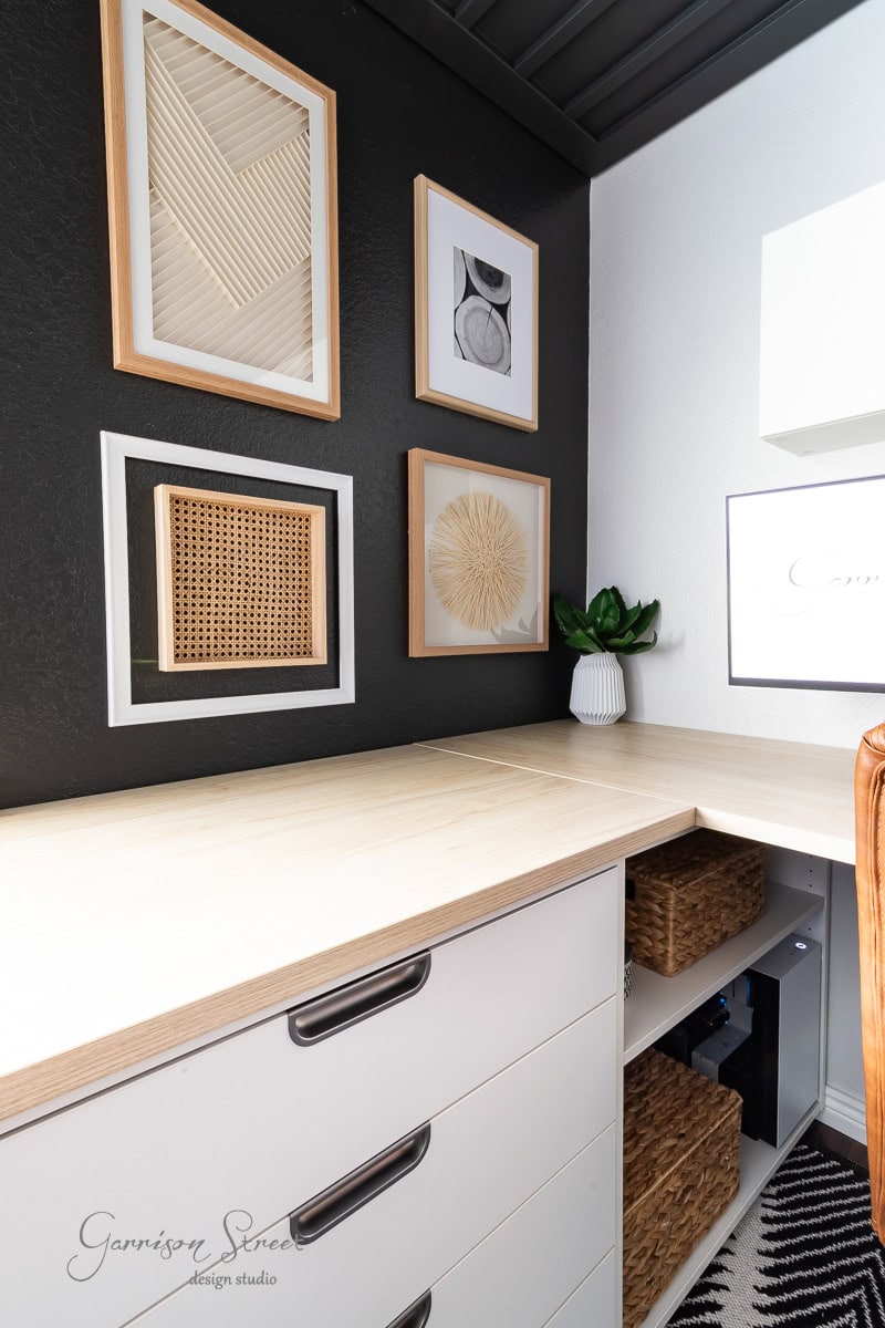 A modern DIY home office corner designed for two features a light wood desk, white drawers, and wicker baskets underneath. Abstract framed artworks adorn the black wall. A small white vase with a plant adds greenery, while a geometric rug is partially visible, completing the cozy workspace.