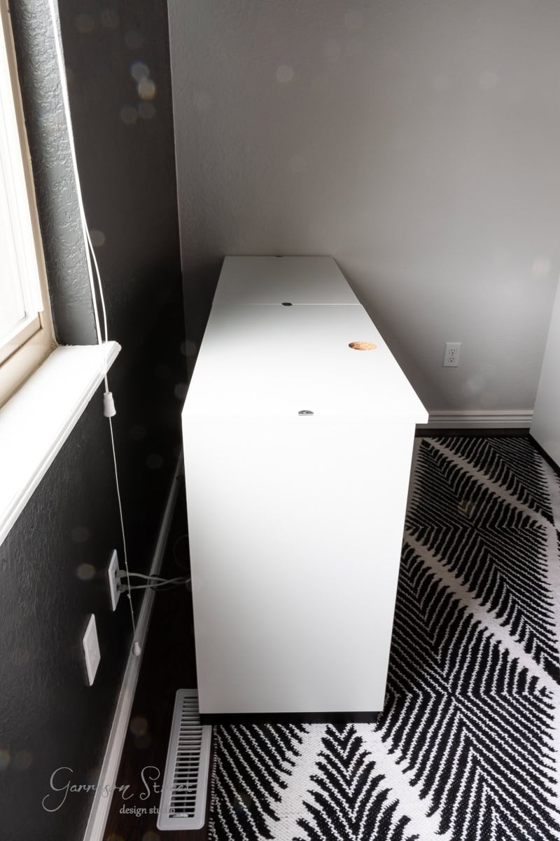 A side view of a minimalist, white desk designed for a DIY home office for two, placed against a dark gray wall. It features a cable management hole, complemented by a patterned black and white rug on the floor. Natural light streams in from the window on the left.