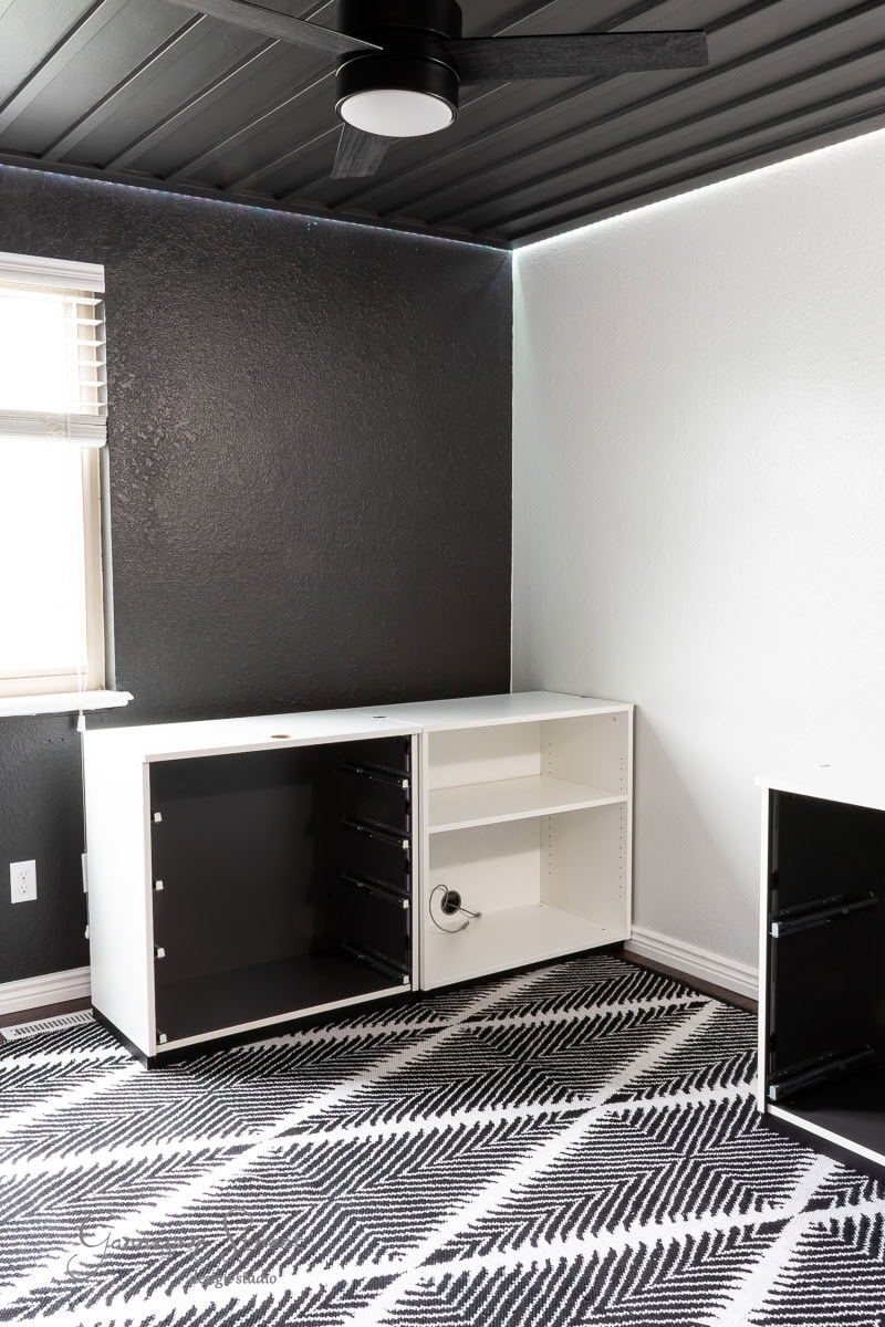 A minimalist DIY home office for two, featuring dark walls and a contrasting white accent wall. It boasts a patterned black and white rug, sleek white cabinets with empty shelves, and a ceiling fan. The space is illuminated by natural light streaming through the window.