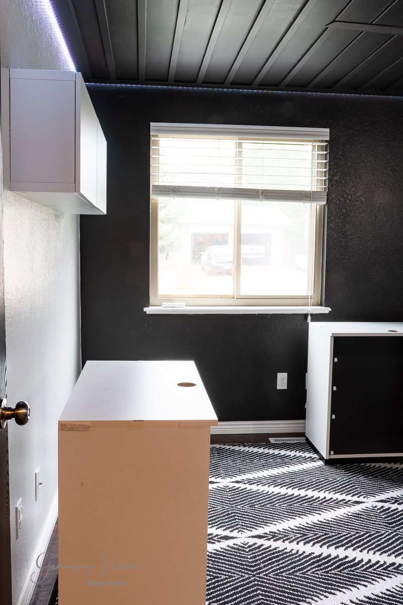 A minimalist DIY home office for two, boasting black walls and ceiling, features a window with blinds. It includes a white desk, a matching wall-mounted cabinet, and a black and white zigzag-patterned rug on the floor. Natural light streams through the window.