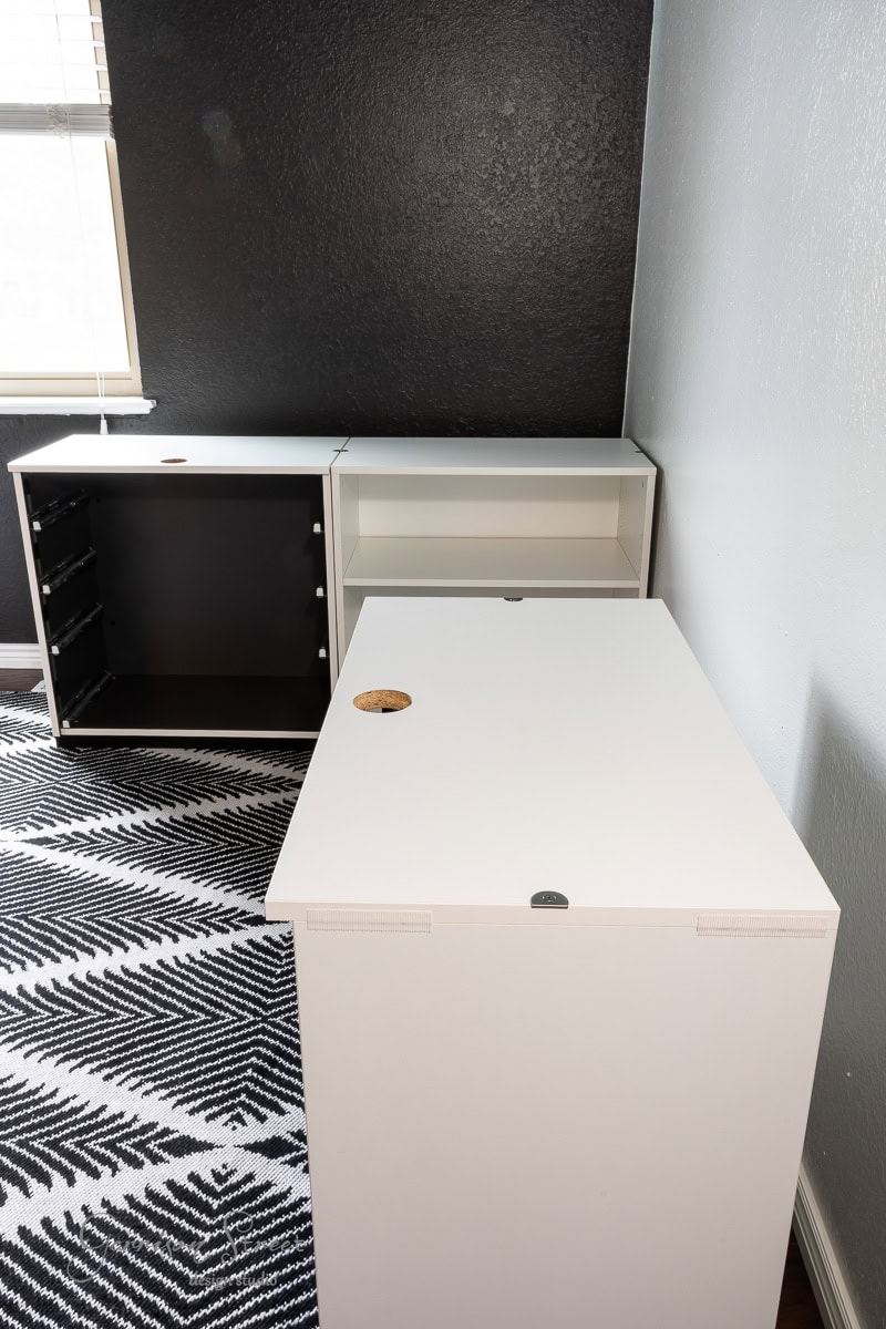 White furniture pieces, including an upright cabinet and a flat piece with a small circular cut-out, set the stage for a stylish DIY home office for two. They sit against a black textured wall, complemented by a black-and-white patterned rug. On the left, a window with blinds offers natural lighting.