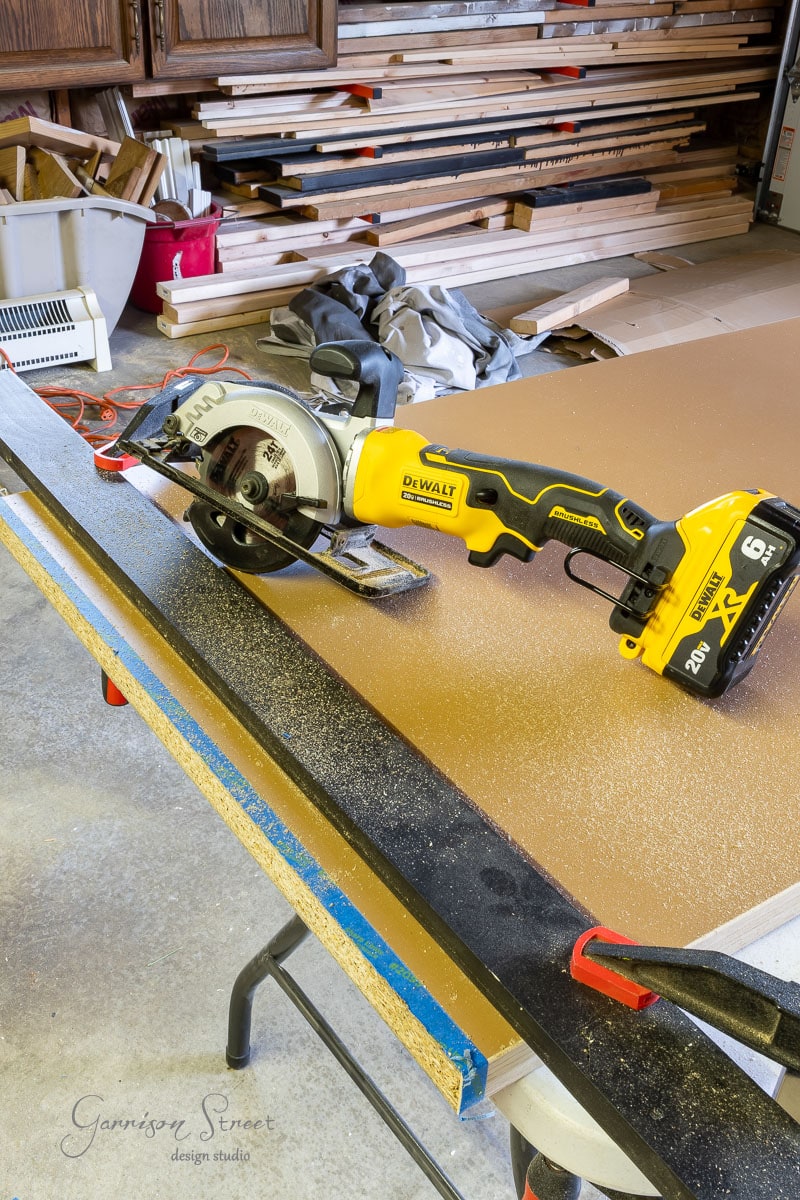 A DeWalt circular saw sits on a workbench dusted with sawdust, embodying the essence of a DIY home office for two. Shelving stacked with planks and tools frames the scene, while markings cover the brown tabletop surface, capturing creativity in action.