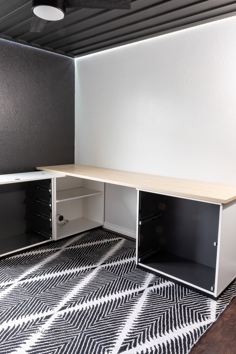 A minimalist room transformed into a DIY home office for two, featuring a black and white zigzag-patterned rug, a light wooden desk with open shelving beneath, dark walls, and a ceiling fan. The space maintains its modern and clean aesthetic.
