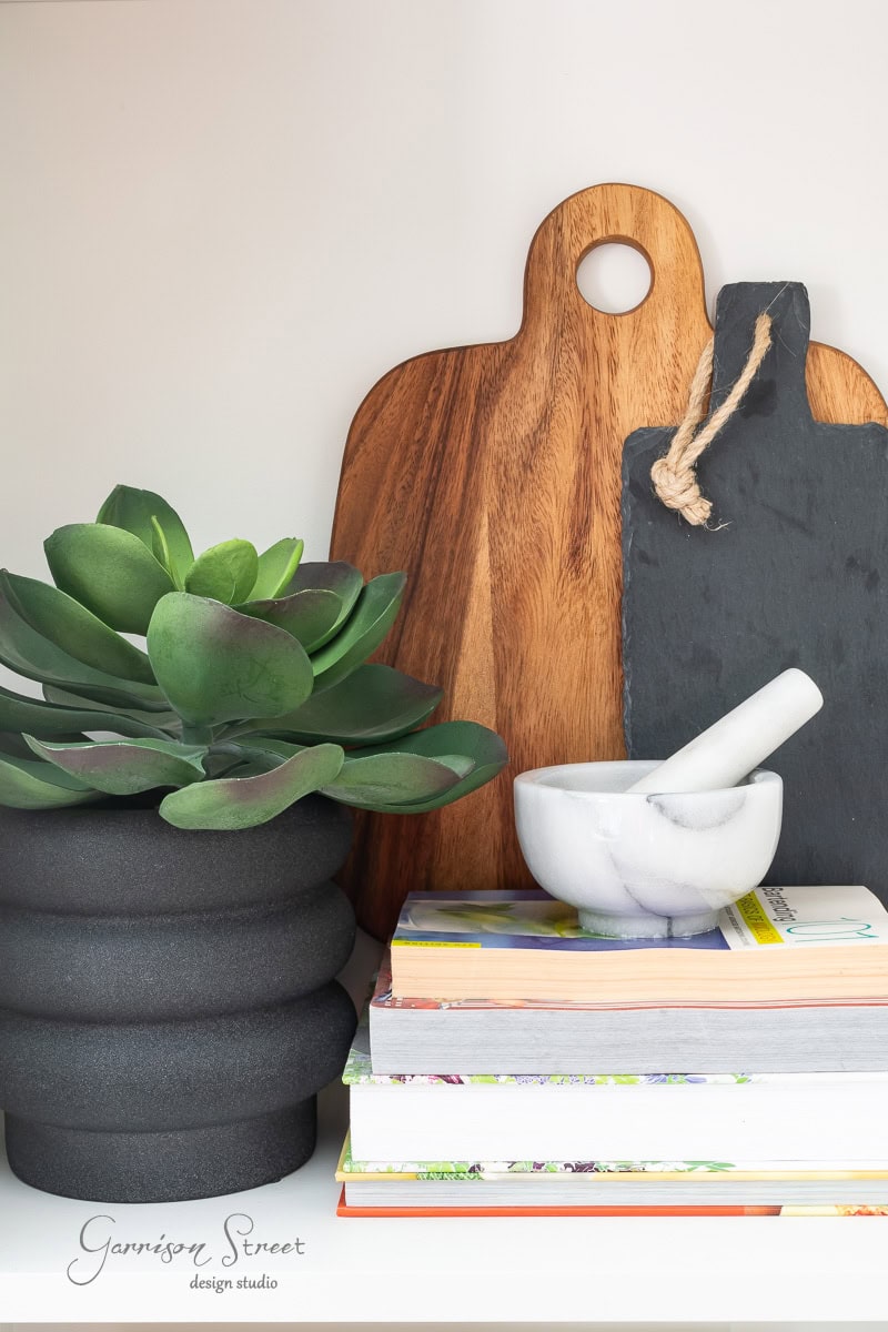 A wooden and slate cutting board stand behind a green succulent in a textured black pot. A marble mortar and pestle sits atop a stack of colorful books, while the text "Garrison Street design studio" graces the lower left corner.