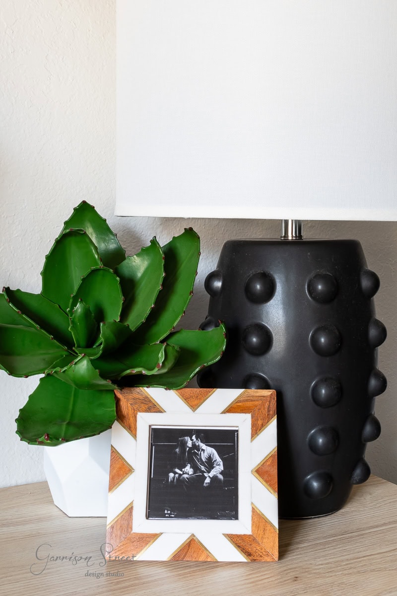 A black lamp with a textured base complements the green faux succulent in a white pot. In front, a photo frame with a zigzag pattern holds a black-and-white picture of a person.