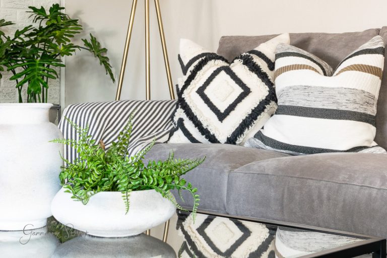 A cozy living room features a gray sofa with decorative black and white patterned pillows. A striped pillow adds contrast. A glass coffee table holds a white ceramic bowl with green ferns, perfect to explore how to decorate with fake plants. A tall floor lamp and a potted plant are in the background.