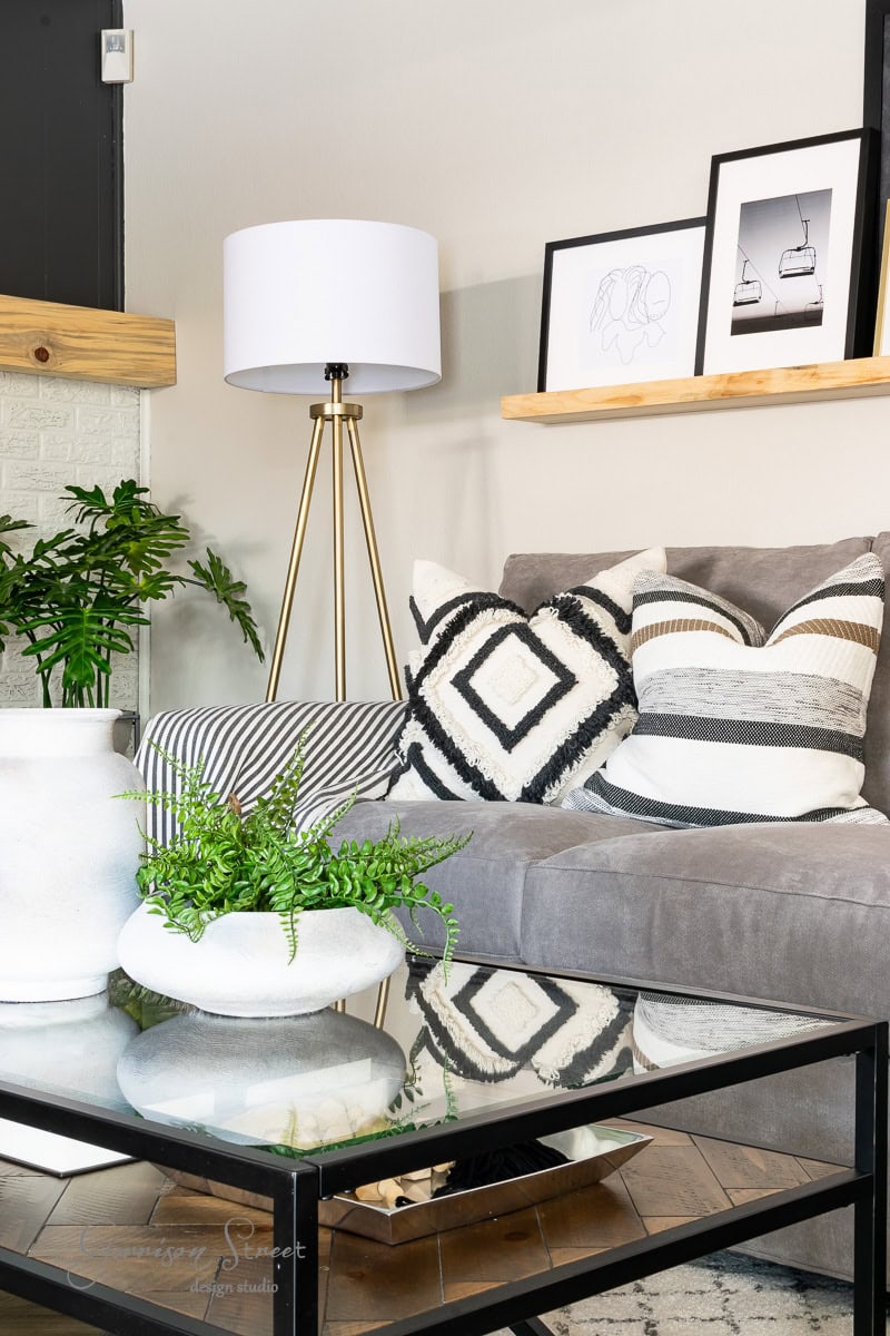 A modern living room features a gray sofa with patterned cushions, a gold floor lamp, and a wall-mounted wooden shelf holding framed art. A glass coffee table adorned with fake plants adds a touch of greenery. Neutral tones and minimalist decor complete the look.