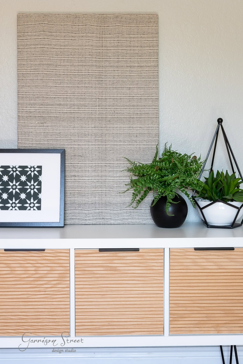 A modern interior with a white and wood cabinet is elegantly accented by a potted fern, a small artificial succulent, and a framed geometric print. A large textured panel is mounted on the wall above the cabinet.