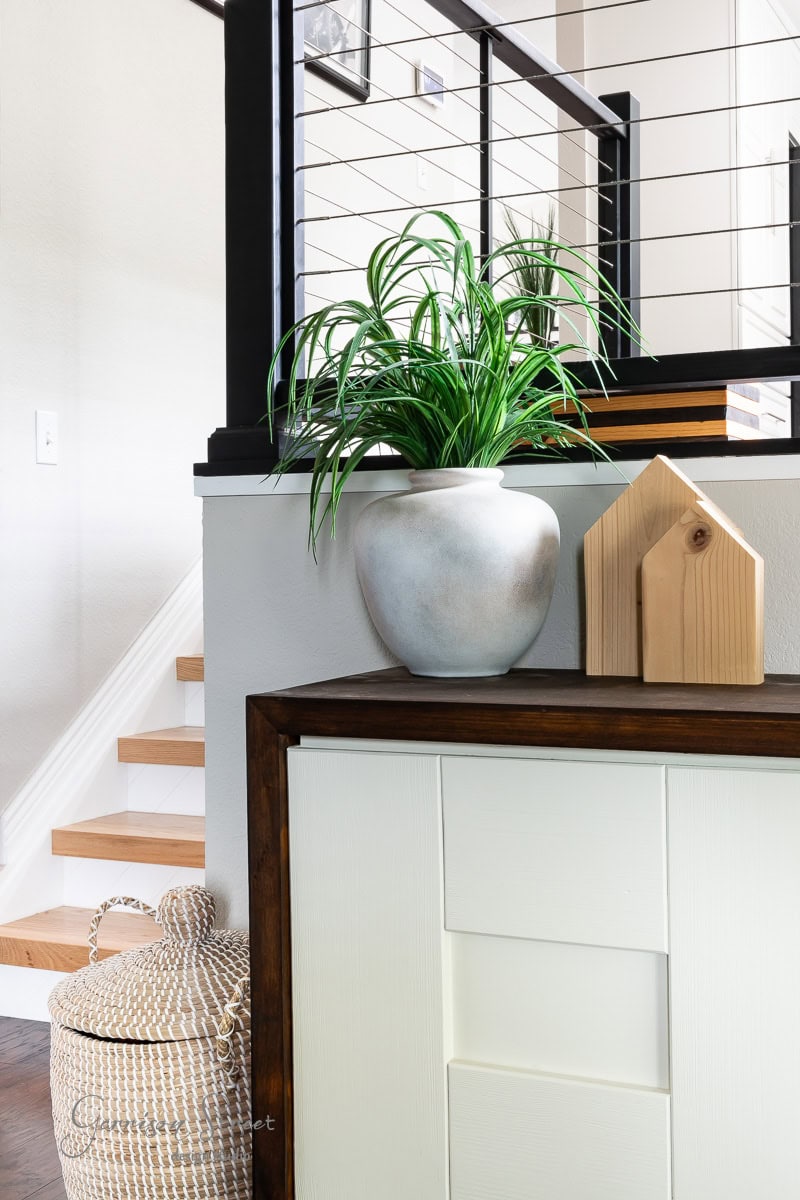 A modern interior features a light gray wall, wooden stairs with a black railing, and a white cabinet with a dark wood top. A textured basket sits on the floor, while a potted plant and wooden decor piece rest on the cabinet.