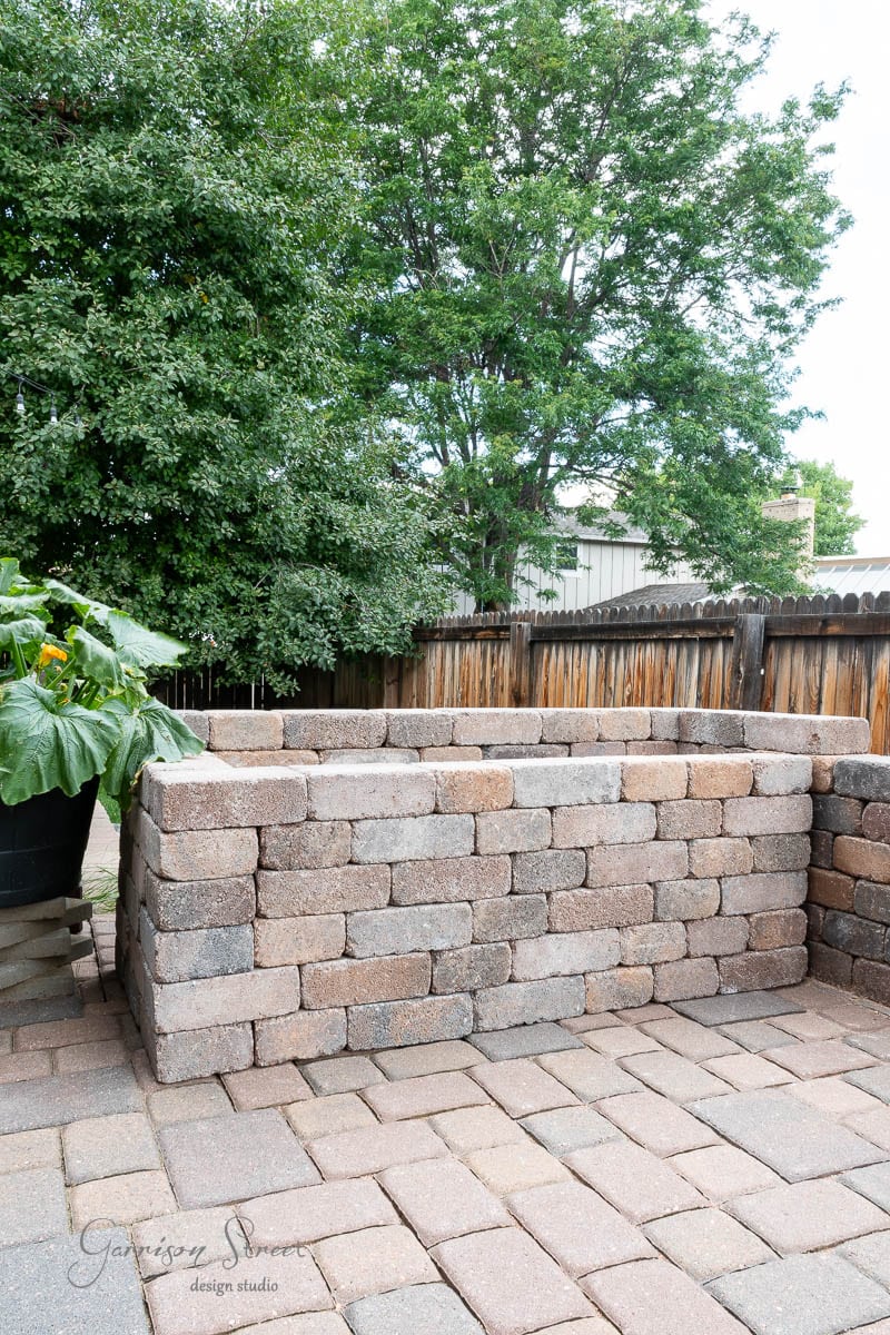 A raised garden bed made of stacked bricks sits proudly on the paved patio, adding charm to the outdoor space. Nearby, a leafy green plant in a pot thrives beside a sleek concrete countertop, and a wooden fence with lush green trees provides a serene backdrop.