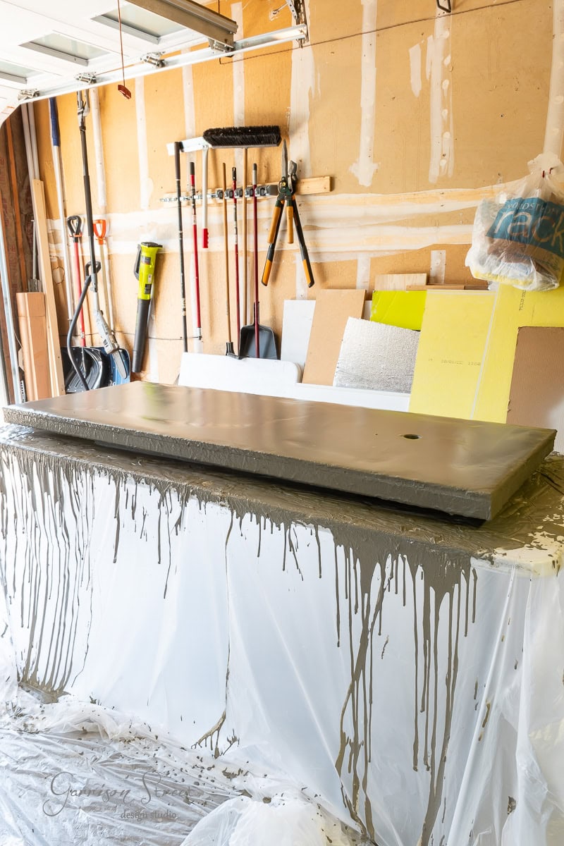 A garage workspace showcases a DIY project with a freshly poured concrete countertop resting on sawhorses, covered in plastic. The walls are lined with various tools, including a broom, rake, and several gardening essentials.