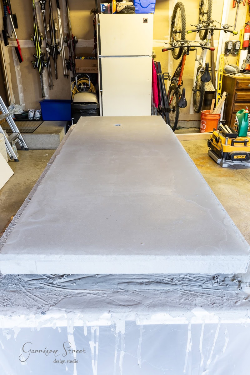 A garage showcases a DIY project, with a large white concrete countertop on a table in the foreground. In the background, you can spot a refrigerator, bicycles, and various tools and storage items neatly arranged, adding to the functional charm of the space.