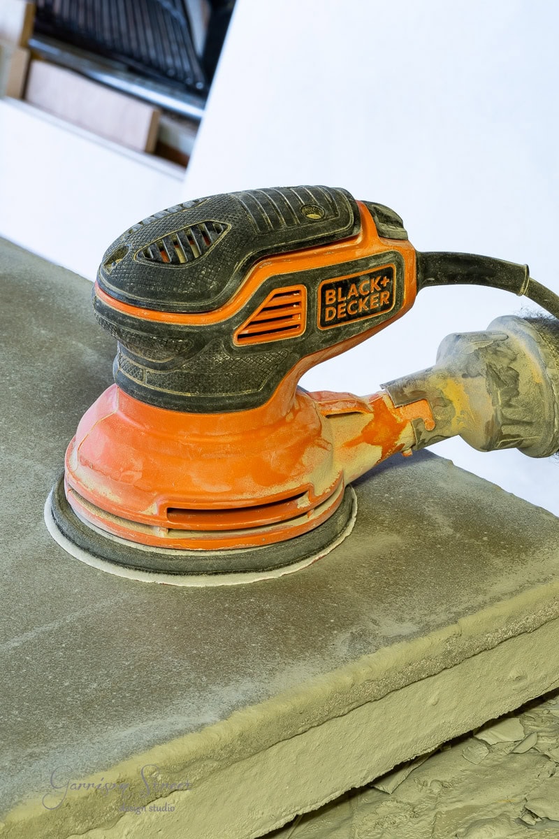 A close-up of a Black & Decker handheld electric sander being used on a flat concrete countertop. Dust swirls around the sander, highlighting its active sanding process. The tool's iconic orange and black design stands out.