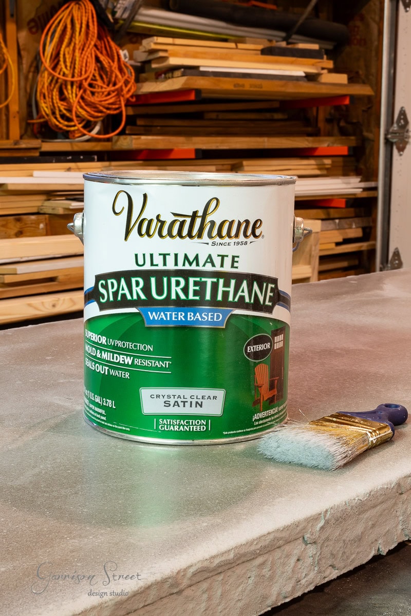 A can of Varathane Ultimate Spar Urethane sits on a workbench alongside a paintbrush. Labeled as water-based with UV protection and resistance to mold and mildew, it's surrounded by tools and wood planks in the background.