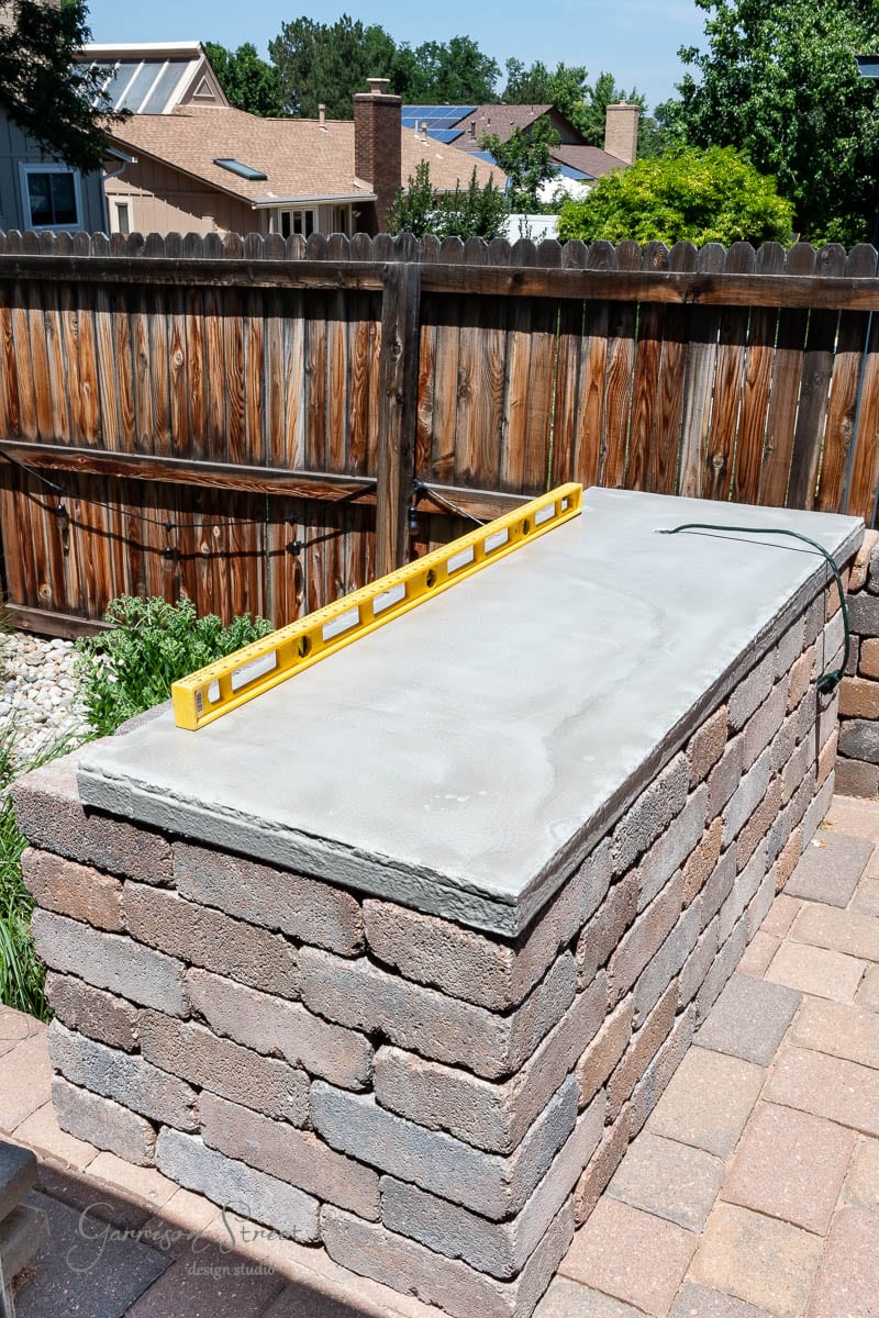 An outdoor kitchen counter is framed by a wooden fence. A yellow level rests on top of the counter, assembled with rectangular stone blocks. In the background, garden plants add a touch of nature to the scene.