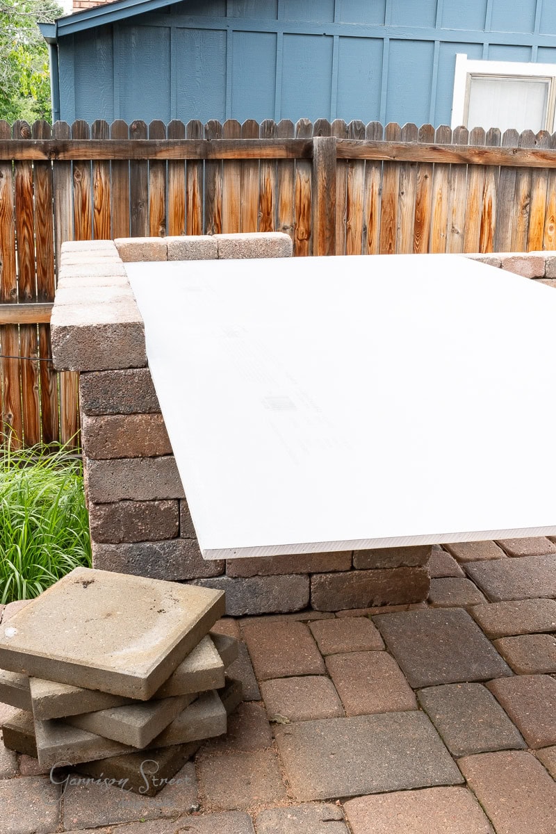 A DIY whiteboard is balanced on stacked cinder blocks in a backyard with a wooden fence and a blue shed in the background. Nearby, additional cinder blocks await their next use on the patio.