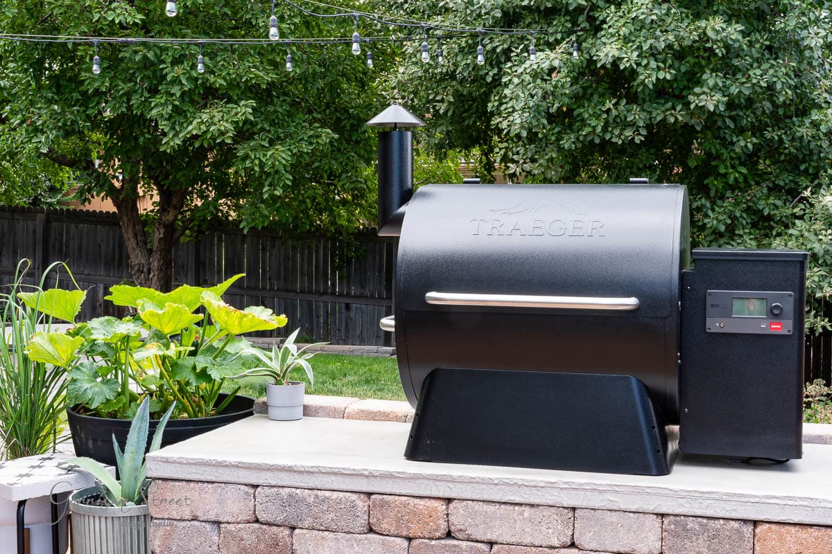 A black Traeger grill sits on a stone outdoor patio with a DIY concrete countertop, surrounded by potted plants. In the background, string lights hang over a lush green garden and a wooden fence.