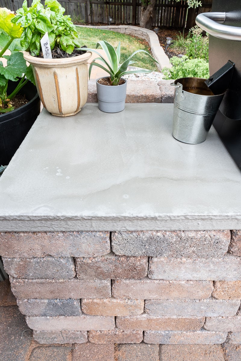 A brick outdoor counter boasts a sleek concrete countertop, complemented by a small potted plant, a green bush, and a metal bucket. In the background, a charming garden with a stone path and wooden fence completes this DIY haven.