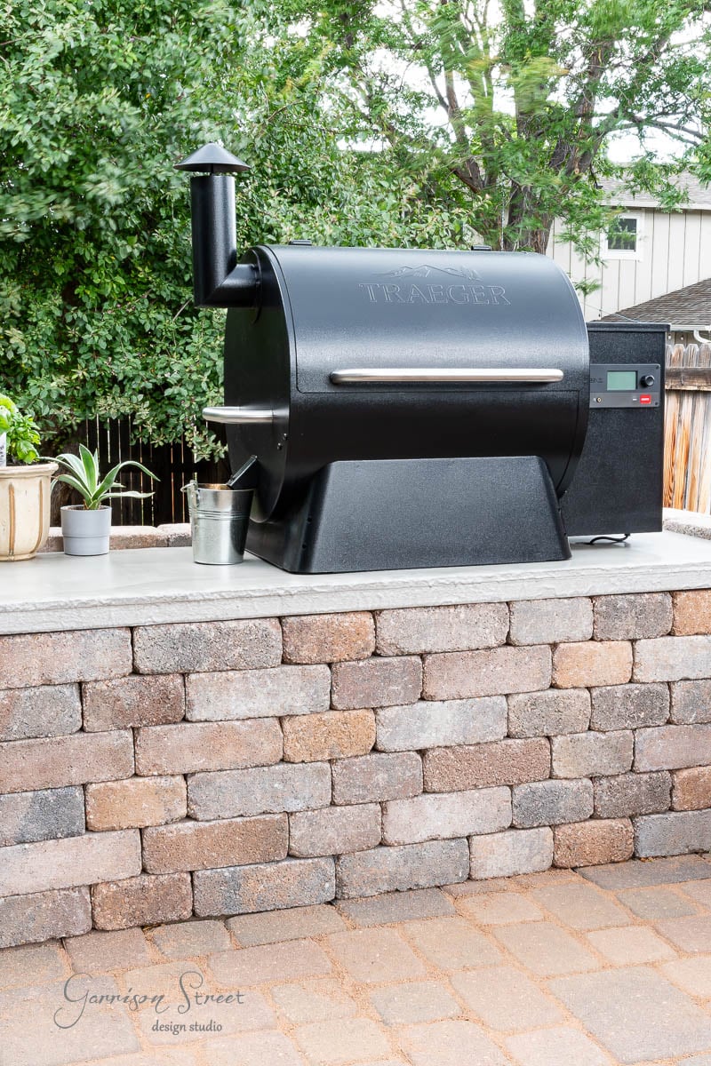 A black Traeger grill rests on a handcrafted concrete countertop, surrounded by potted plants and a small bucket. In the background, a fence mingles with lush trees.