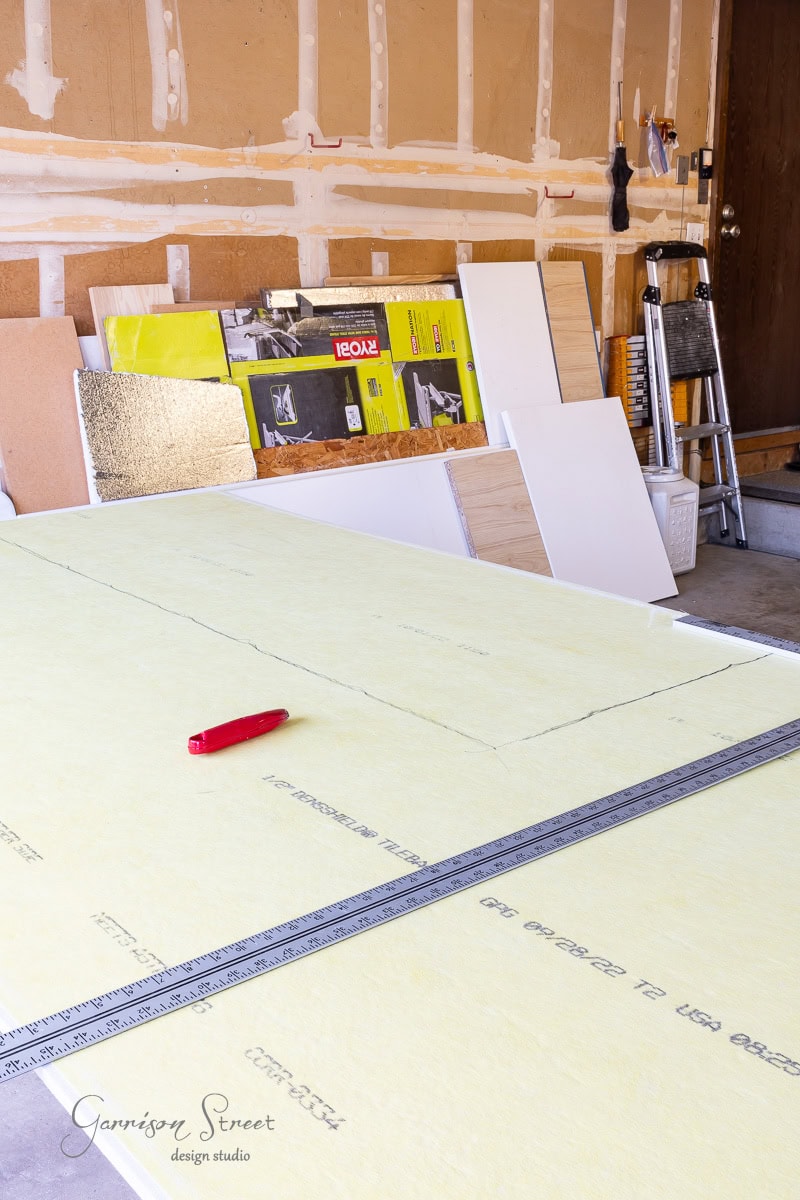A garage workspace brimming with construction materials features a large sheet of yellow insulation board on a table, alongside a metal ruler and red utility knife. Various boards and tools are stacked against the wall.