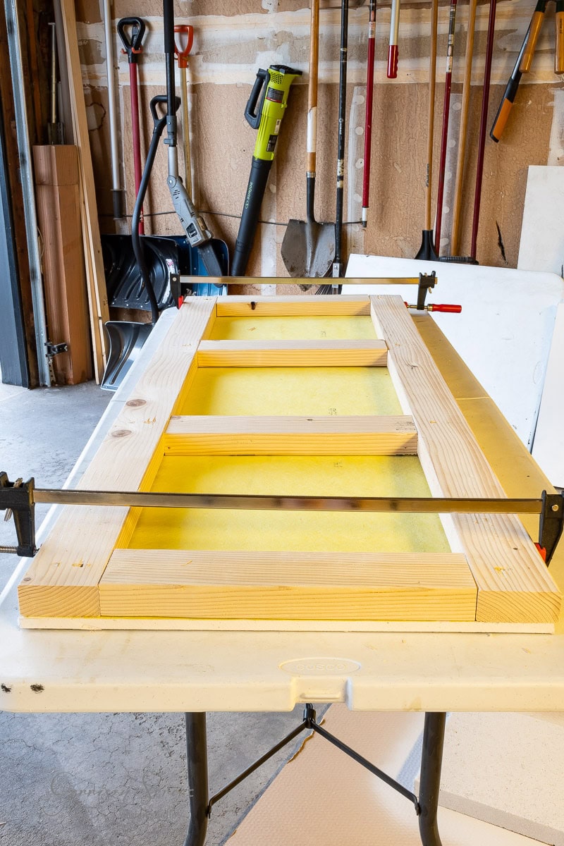 A wooden frame in the process of being assembled on a workbench, held by clamps. Various tools, including rakes and a leaf blower, hang on the wall in the background of a garage or workshop.