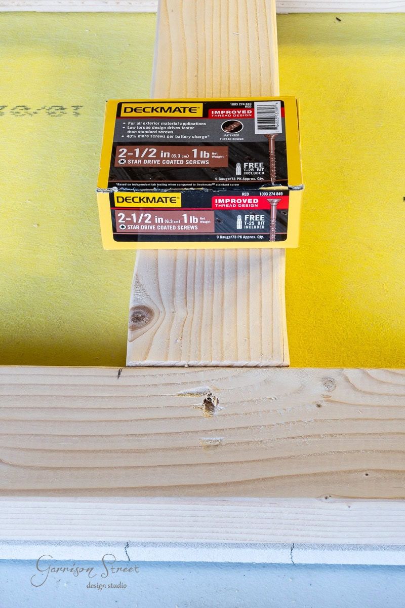 A box of Deckmate 2-1/2 inch star drive coated screws sits on a piece of wood. The wood rests on a yellow surface, creating a construction scene. The text "Garrison Street Design Studio" is visible at the bottom left.
