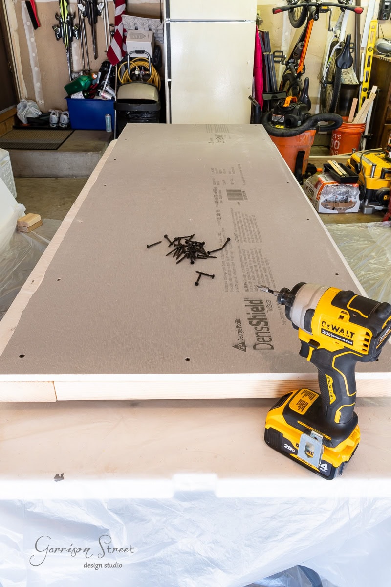 A workbench in a garage is set up for a project, featuring a DeWalt drill beside a pile of screws and a board ready for assembly. In the background, tools and equipment are visible.