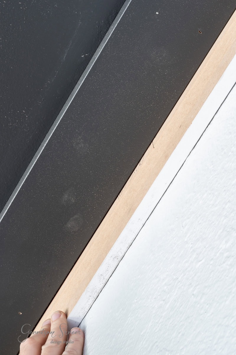 A hand holds a piece of unfinished wooden trim against a wall with contrasting colors, one side dark gray and the other light blue. The trim, reminiscent of a diy wood plank ceiling project, elegantly defines the boundary between the two sections.
