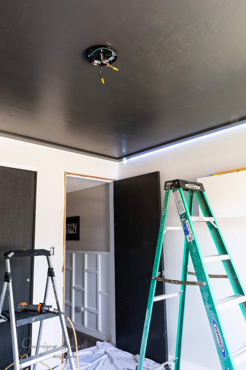The room under renovation features a striking black ceiling and a green ladder. Wires dangle from above, hinting at the next phase of this DIY project. Walls are painted white, and there's an opening to another room with tools scattered across a plastic-covered floor.
