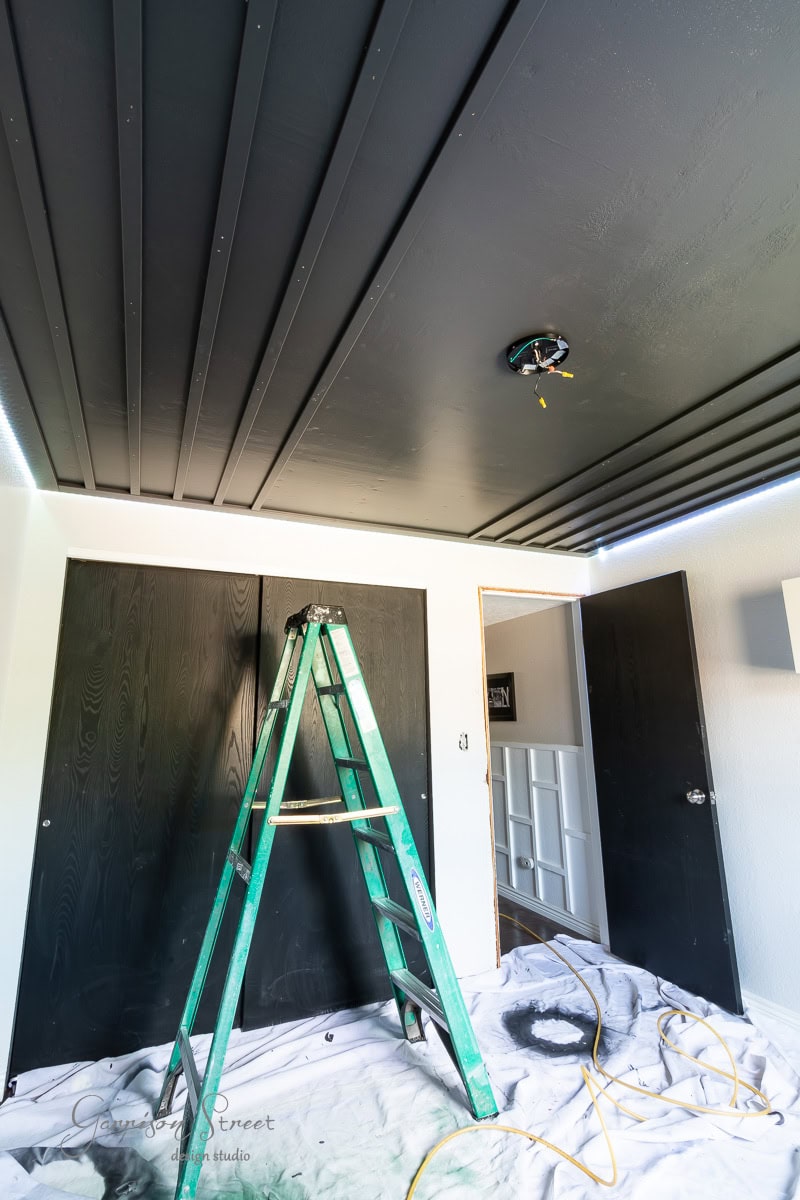 A room under renovation features a partially open door and a black painted ceiling with exposed wooden beams, perfect for a DIY wood plank ceiling project. A green ladder stands beneath an unfinished light fixture, while the floor is shielded by a white sheet, scattered with tools.