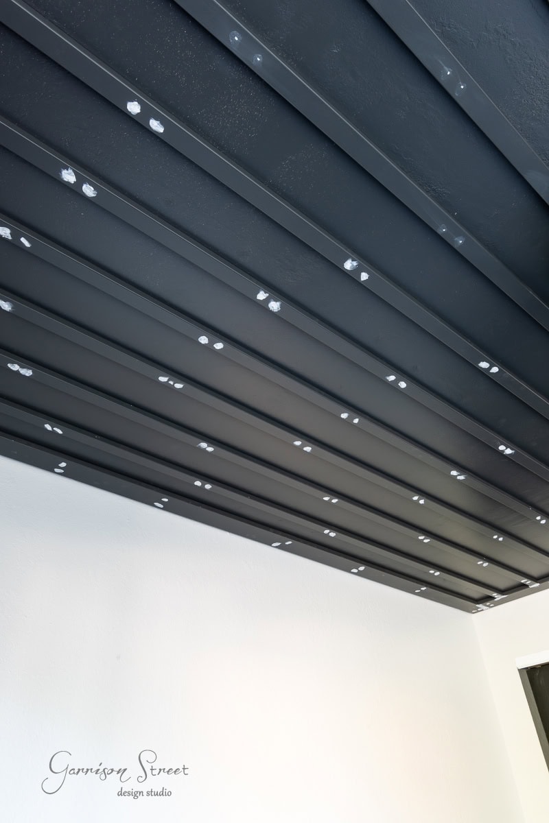 The image showcases a modern diy wood plank ceiling with black beams and exposed screws. The evenly spaced beams create a sleek, industrial vibe. A section of white wall peeks beneath the ceiling, while "Garrison Street Design Studio" is elegantly displayed at the bottom.