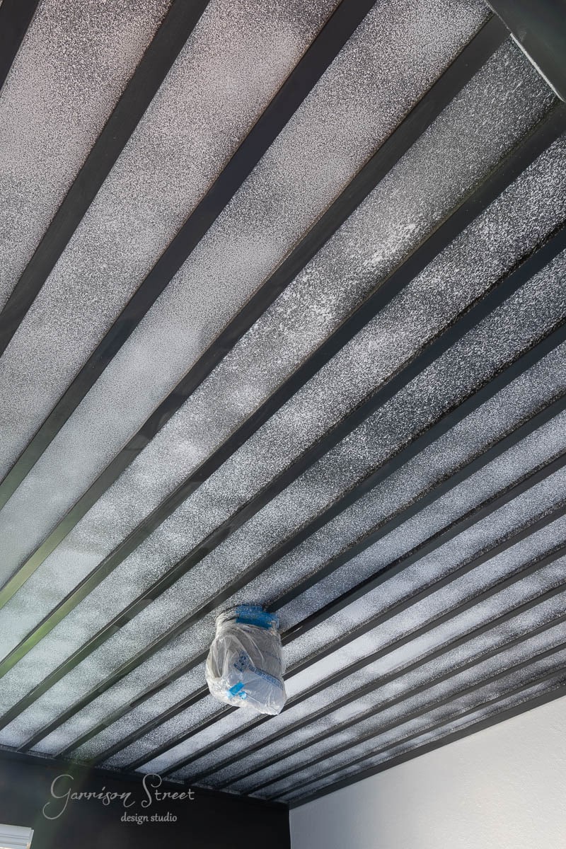 A ceiling featuring contrasting dark beams and a light textured finish evokes a DIY wood plank ceiling charm. A light fixture, wrapped for protection, hangs in the foreground. The design beautifully combines modern and rustic elements.